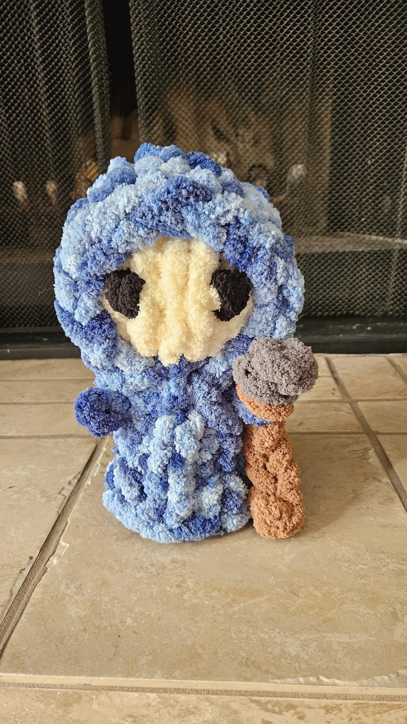 May include: A handmade crochet doll in shades of blue and white, with a cream-colored face and dark brown eyes. The doll is holding a brown and gray object, possibly a staff or a toy. The doll is standing on a tile floor.