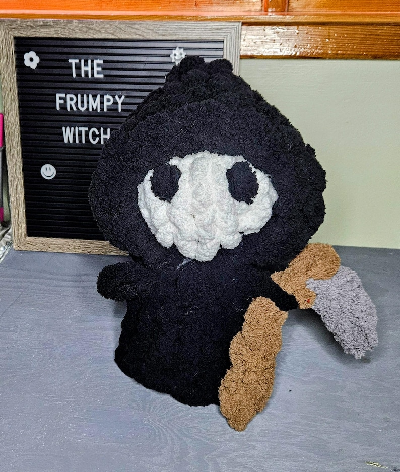 May include: A handmade black plush Grim Reaper doll with a white skull face and a brown and gray scythe. The doll is next to a framed letter board that reads "THE FRUMPY WITCH".