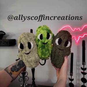 May include: Three plush, smiling, alien-like creatures in shades of green and brown. Each has large white eyes and a stitched black smile. The text "@allyscoffincreations" is above the creatures. The background includes a neon bat and black candles.