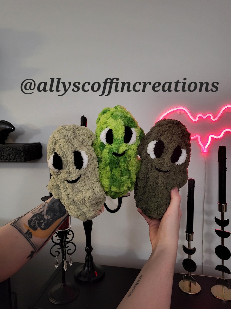 May include: Three plush, smiling, alien-like creatures in shades of green and brown. Each has large white eyes and a stitched black smile. The text "@allyscoffincreations" is above the creatures. The background includes a neon bat and black candles.