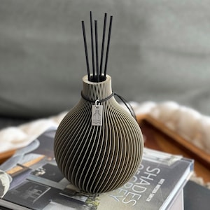 Può includere: Un diffusore in ceramica grigia con un design a coste e bastoncini diffusori neri. La bottiglia ha un'etichetta argentata con il testo "ICONIC HOME".