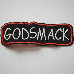 May include: Embroidered patch featuring the word "GODSMACK" in white, with a black background and a red border. The patch has a rectangular shape with a wavy edge.