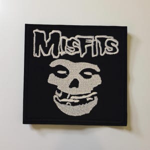 Op de afbeelding: Een zwart vierkant embleem met het woord "MISFITS" in witte letters boven een witte schedelafbeelding. De schedel heeft een brede grijns. De patch heeft een zwarte rand en is waarschijnlijk voor kleding.