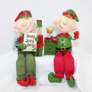 May include: Two Christmas elf figurines. One holds a sign that reads "Holly Jolly Day!" and the other holds a gift box that reads "Golly Be Jolly". Both elves wear red and green outfits and hats.