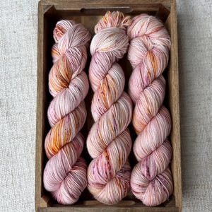 Könnte beinhalten: Drei Stränge handgefärbter Wolle in einer Holzkiste. Das Garn ist eine Mischung aus rosa, pfirsichfarbenen und orangenen Farbtönen, die einen weichen, bunten Farbeffekt erzeugt. Die Holzkiste hat eine natürliche Oberfläche und bietet einen rustikalen Kontrast.