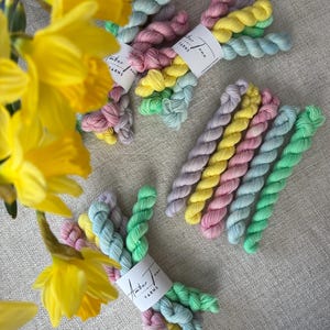 Può includere: Sono presentati diversi gomitoli di filato dai colori pastello, rosa, giallo, verde e blu. Ogni gomitolo è avvolto con un'etichetta bianca con la scritta "Amber Tone Yarns". I narcisi gialli sono sullo sfondo.