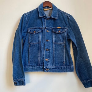 May include: A blue denim jacket with a button closure and two front pockets. The jacket has a brown leather label with the text "Wrangler" on the left chest.