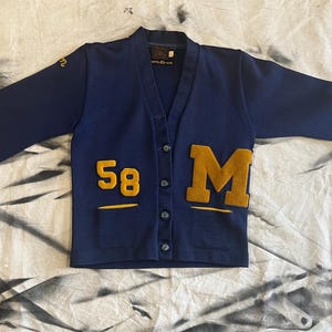 May include: A navy blue cardigan with a gold '58' and 'M' on the front. The cardigan has a button-up front and long sleeves.