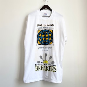 以下が含まれることがあります： 地球儀、テニスラケット、そして「WORLD TOUR」と「BISHOP PARK BREAKERS」の文字が描かれたグラフィックデザインの白いTシャツ。 シャツは黒いハンガーにかかっています。
