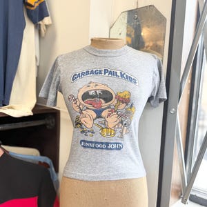 May include: A grey t-shirt with a Garbage Pail Kids graphic. The design shows a cartoon character with an open mouth, surrounded by food. The text "GARBAGE PAIL KIDS" and "JUNKFOOD JOHN" are also visible on the shirt.