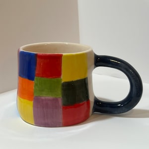 May include: A white ceramic mug with a geometric pattern of colorful squares in shades of blue, red, yellow, green, purple, and black. The mug has a dark blue handle.