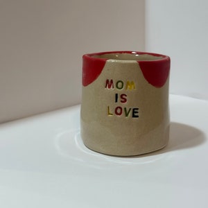 Puede incluir: Una taza pequeña de cerámica beige con letras rojas, amarillas y verdes que dicen "MOM IS LOVE".