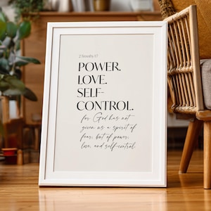 May include: A framed print with the words "POWER. LOVE. SELF-CONTROL." in a minimalist design. The text is in black against a cream background, with the Bible verse 2 Timothy 1:7 at the top. The frame is white.