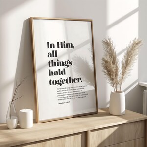 May include: A framed print with the text "In Him, all things hold together" in black font. The print is displayed on a light wood cabinet, alongside a vase with dried plants and a small candle.
