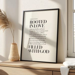 May include: A framed print with the text "ROOTED IN LOVE" and "FILLED WITH GOD" in a black frame. The print is on a cream-colored background with a Bible verse. A decorative vase with dried plants sits on a wooden surface.