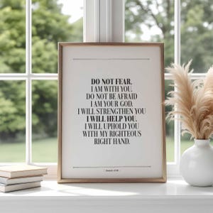 May include: A framed print with a light brown wooden frame. The print features black text on a white background, with the words "DO NOT FEAR, I AM WITH YOU." The print is displayed on a white windowsill with books and a vase.