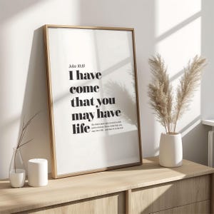 May include: A framed print with the text "I have come that you may have life" in a minimalist design. The print is in a light wood frame and is displayed on a wooden cabinet. A vase with dried plants is next to the print.