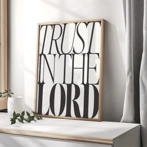 Può includere: Una stampa artistica incorniciata con la scritta "TRUST IN THE LORD" in grandi lettere nere su sfondo bianco. La cornice è in legno chiaro. La stampa è esposta su una superficie bianca, adatta all'arredamento.