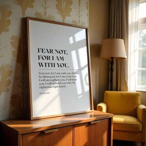Pode incluir: Uma impressão emoldurada com o texto "FEAR NOT, FOR I AM WITH YOU." A impressão está em uma moldura de madeira e é exibida em um armário de madeira. O quarto tem uma poltrona amarela e uma lâmpada.