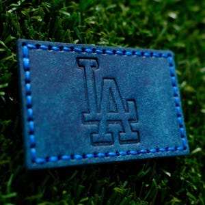 May include: A blue leather patch with white stitching around the edges. The patch features the letters "LA" in a stylized font.