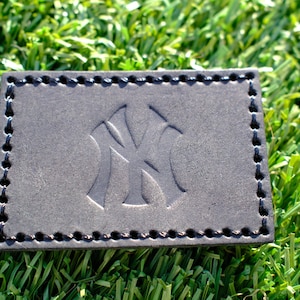 May include: A rectangular, dark gray leather patch with the embossed interlocking "NY" logo, stitched around the edges with black thread. The patch is set against a backdrop of green artificial turf.