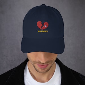 Puede incluir: Una gorra de béisbol azul marino con un corazón roto rojo bordado y una lágrima azul. El texto "HEART BREAKER" está bordado en amarillo debajo del corazón.