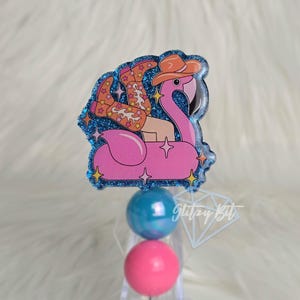 May include: A badge featuring a pink flamingo floatie with a person wearing cowboy boots and a hat. The badge has a blue glitter border with star accents. Below are two round beads, one blue and one pink.