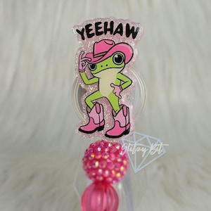 May include: A badge reel featuring a glittery acrylic topper with a green frog wearing a pink cowboy hat and pink cowboy boots. The text YEEHAW is printed in black above the frog. Below the topper are two pink beads, one textured and one smooth.