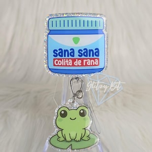 May include: A badge reel with a blue and white jar design, featuring the text "sana sana colita de rana." A green frog charm is attached below. The badge reel has a glittery border.