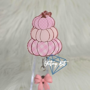 May include: A pink pumpkin-shaped badge reel with a glittery outline. The pumpkin design features a mix of polka dots and gingham patterns. A pink bow and a blue diamond shape are also visible.