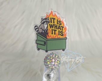 May include: A badge reel with a cartoon raccoon sitting on a green dumpster engulfed in flames. The text "IT IS WHAT IT IS" is above the dumpster. The reel has a sparkly, iridescent design.