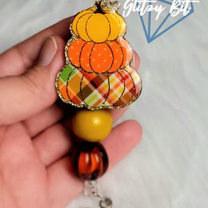 May include: A fall-themed badge reel featuring a stacked pumpkin design. The top pumpkin is orange with gold glitter trim, the middle pumpkin is orange with white polka dots, and the bottom pumpkin has a plaid pattern. The reel includes yellow and orange beads.