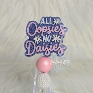 May include: A badge reel with the phrase "ALL Oopsies NO Daisies" in pink and white lettering on a blue glitter background. The reel has a pink bead and a white pearl bead at the bottom.