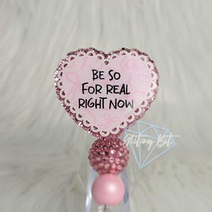 May include: A pink heart-shaped badge reel with the phrase "BE SO FOR REAL RIGHT NOW" in black text. The badge reel has a glittery border and a pink beaded accent.