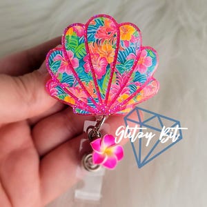 May include: A seashell-shaped badge reel with a tropical floral pattern in pink, orange, yellow, and teal. The badge reel has a pink glitter border and a small pink flower charm. The text "Glitzy Bit" is visible.