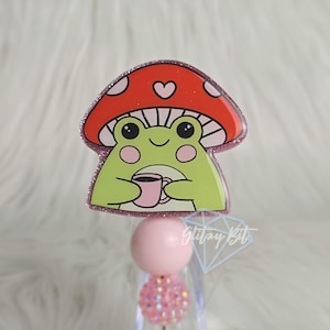 May include: A whimsical badge featuring a green frog under a red mushroom cap with white spots and a heart. The frog is holding a pink cup. The badge has a pink glitter border and a pink and crystal bead accent.