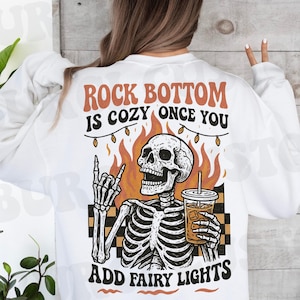 May include: White sweatshirt with a graphic of a skeleton holding a drink, flames, and the text "ROCK BOTTOM IS COZY ONCE YOU ADD FAIRY LIGHTS" in orange and black. The skeleton is giving a rock and roll hand gesture.