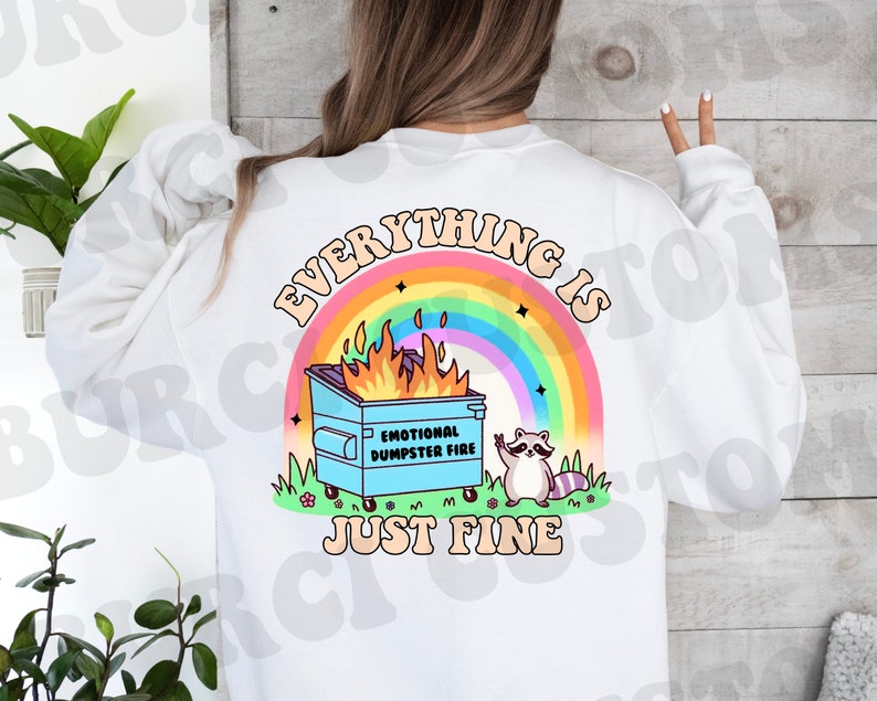 K&ouml;nnte beinhalten: Wei&szlig;es Sweatshirt mit einem Regenbogen und einem blauen M&uuml;llcontainer in Flammen mit dem Text "Alles ist gut" und einem Waschb&auml;ren mit einem Peace-Zeichen.