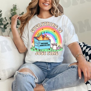 K&ouml;nnte beinhalten: Ein wei&szlig;es T-Shirt mit dem Aufdruck "Everything is just fine". Der Text ist von einem Regenbogen und einem Cartoon-Bild eines M&uuml;lltonnenfeuers mit einem Waschb&auml;ren daneben umgeben. Auf der M&uuml;lltonne steht der Text "Emotional Dumpster Fire".