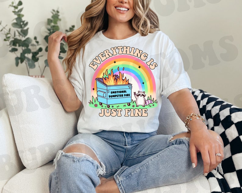 K&ouml;nnte beinhalten: Ein wei&szlig;es T-Shirt mit dem Aufdruck "Everything is just fine". Der Text ist von einem Regenbogen und einem Cartoon-Bild eines M&uuml;lltonnenfeuers mit einem Waschb&auml;ren daneben umgeben. Auf der M&uuml;lltonne steht der Text "Emotional Dumpster Fire".