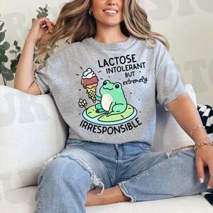 Puede incluir: Una camiseta gris con una rana verde que lleva un sombrero y sostiene un cono de helado. El texto en la camiseta dice "Lactose Intolerant But Extremely Irresponsible".
