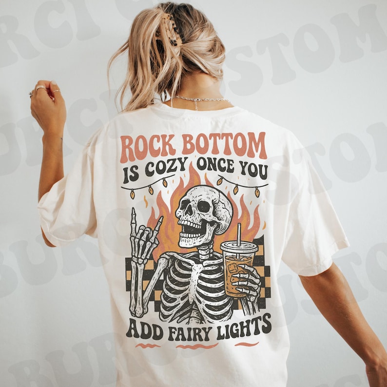 May include: Cream-colored t-shirt with a graphic of a skeleton holding a drink, flames, and the text "Rock Bottom Is Cozy Once You Add Fairy Lights." The skeleton is giving a rock and roll hand gesture.