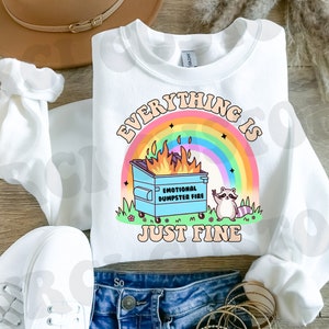 K&ouml;nnte beinhalten: Wei&szlig;es Sweatshirt mit einem Regenbogen und einem blauen M&uuml;llcontainerbrand mit dem Text "Everything is Just Fine" und einem Waschb&auml;ren mit einem Peace-Zeichen.
