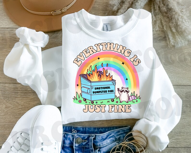 K&ouml;nnte beinhalten: Wei&szlig;es Sweatshirt mit einem Regenbogen und einem blauen M&uuml;llcontainerbrand mit dem Text "Everything is Just Fine" und einem Waschb&auml;ren mit einem Peace-Zeichen.