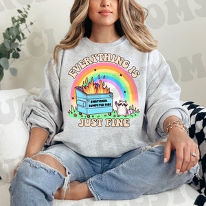 K&ouml;nnte beinhalten: Ein graues Sweatshirt mit dem Text "Everything is just fine" und einem Cartoon-Bild eines Waschb&auml;ren, der neben einem M&uuml;lltonnenfeuer steht, mit dem Text "Emotional Dumpster Fire".