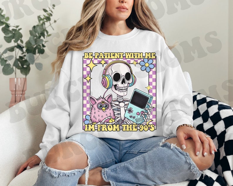 May include: White sweatshirt with a graphic of a skeleton wearing headphones and holding a video game controller. The text "Be patient with me I'm from the 90's" is printed on the sweatshirt.