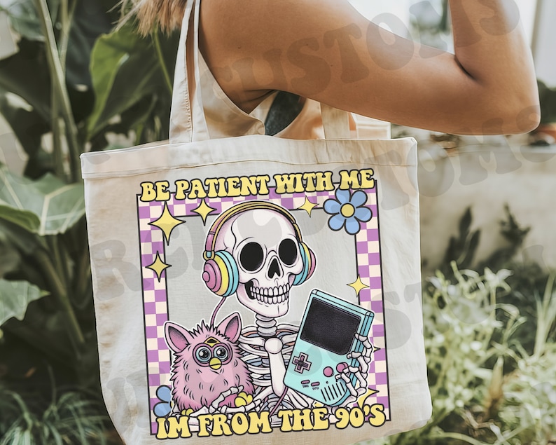 May include: A white tote bag with a pink, blue, and yellow graphic of a skeleton wearing headphones and holding a video game. The text on the bag reads "Be patient with me, I'm from the 90's".