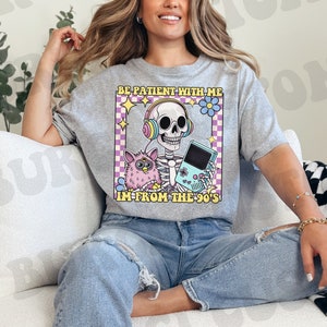 May include: A gray t-shirt with a graphic of a skeleton wearing headphones and holding a video game console. The graphic also includes a fuzzy pink toy and the text "Be patient with me, I'm from the 90's".