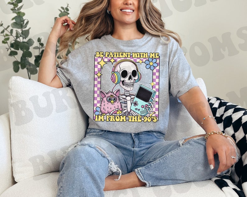 May include: A gray t-shirt with a graphic of a skeleton wearing headphones and holding a video game console. The graphic also includes a fuzzy pink toy and the text "Be patient with me, I'm from the 90's".