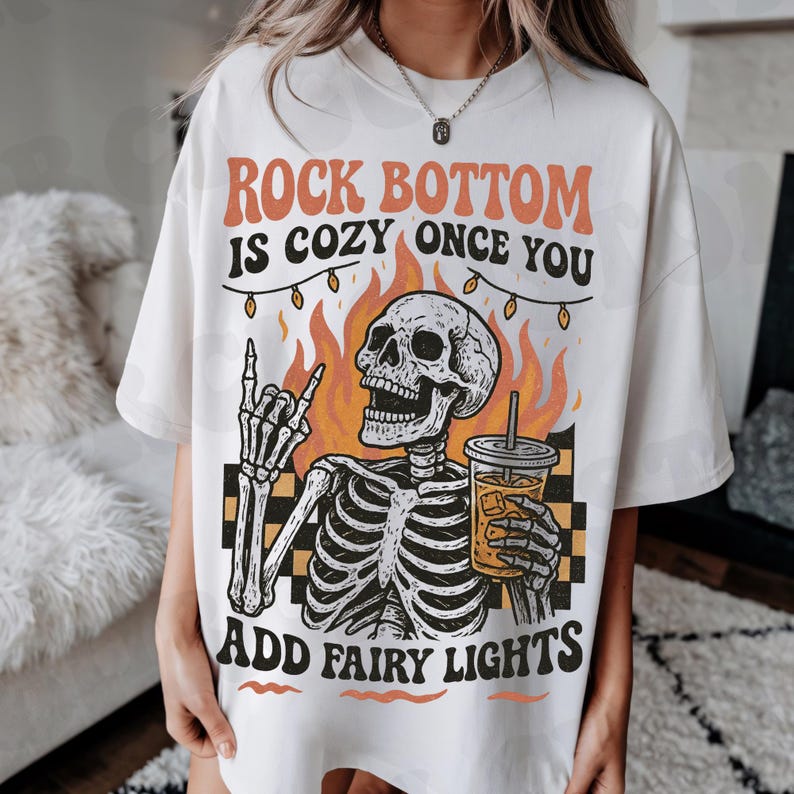 May include: Cream-colored t-shirt with the text "ROCK BOTTOM IS COZY ONCE YOU ADD FAIRY LIGHTS." The graphic features a skeleton with a coffee drink, flames, and a string of lights. The skeleton is giving a rock and roll hand gesture.